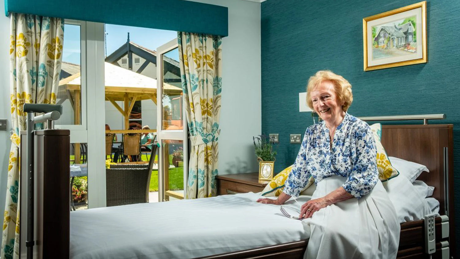 An older lady sat on a Opera profiling bed in a stylish care home.
